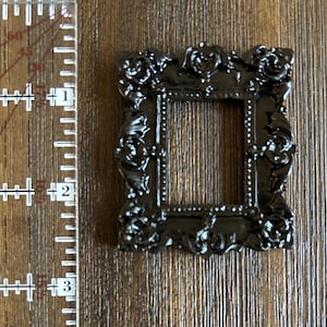 May include: A small, ornate black picture frame with intricate floral and scroll detailing. The frame is square, with a central opening. The frame measures approximately 1.5 inches in height and width.