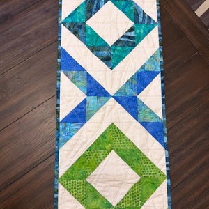 May include: A rectangular quilted table runner featuring a geometric pattern of diamonds and triangles. The design incorporates shades of teal, blue, and green fabric pieces contrasted against off-white fabric, with visible machine quilting stitches.
