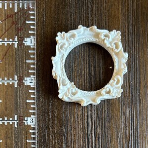 May include: A small, ornate, white resin frame with a circular opening. The frame has a detailed, vintage design with scrolling patterns. The frame is approximately 2 inches in diameter and sits on a wooden surface.