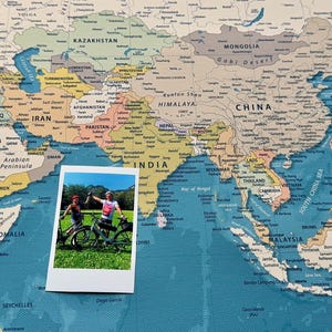 Peut inclure: Une photographie rectangulaire montrant deux personnes debout avec des vélos sur un terrain herbeux, posée sur une grande carte du monde détaillée. La carte affiche divers pays, océans et étiquettes géographiques dans une palette de couleurs douces.