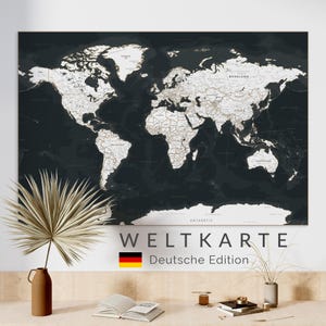 May include: A large, dark world map with white continents and country names. The map is labeled "WELTKARTE Deutsche Edition" in white text. The German flag is displayed below the text. The map is mounted on a white wall.
