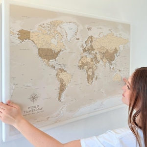 May include: A framed world travel map in neutral tones, with a compass rose and the text "WORLD TRAVEL MAP". The map is being held up against a white wall.