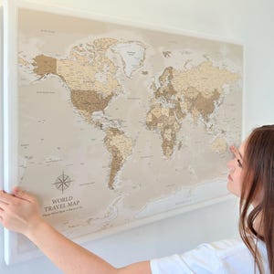 Puede incluir: Un mapa del mundo enmarcado con una paleta de colores neutros, con una rosa de los vientos y el texto "WORLD TRAVEL MAP". El mapa se muestra contra una pared blanca.