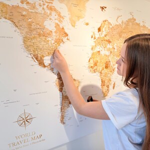 May include: A large wooden world travel map with a light brown finish is mounted on a white wall. The map features detailed geographical outlines and text. A person is placing a pin on the map. The map includes the text "WORLD TRAVEL MAP".