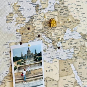 Puede incluir: Un mapa de lienzo beige de Europa y el norte de África con texto negro para nombres de países y ciudades. Varias chinchetas redondas blancas, marrones y doradas están insertadas. Una chincheta dorada con forma de casa está sobre Polonia.