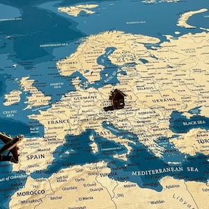 May include: A vintage-style map of Europe featuring a blue ocean and beige landmasses. A small brown wooden airplane figurine is positioned over the Iberian Peninsula, and a small brown wooden house figurine is placed over Central Europe.