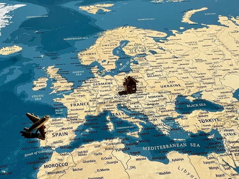 May include: A vintage-style map of Europe featuring a blue ocean and beige landmasses. A small brown wooden airplane figurine is positioned over the Iberian Peninsula, and a small brown wooden house figurine is placed over Central Europe.
