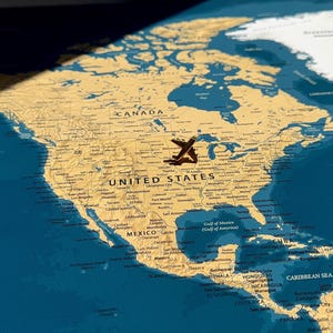 May include: A wall map of North America featuring a tan landmass against a deep blue ocean background. A small metal airplane pin is positioned over the United States, while another pin is visible in the lower right corner near Venezuela.