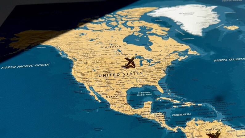 May include: A wall map of North America featuring a tan landmass against a deep blue ocean background. A small metal airplane pin is positioned over the United States, while another pin is visible in the lower right corner near Venezuela.
