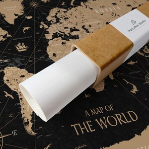 Könnte beinhalten: Eine gerollte Weltkarte in einer braunen Pappröhre mit weißem Etikett. Die Karte ist schwarz mit goldfarbenen Kontinenten und nautischen Illustrationen. Der Text "A MAP OF THE WORLD" ist sichtbar.