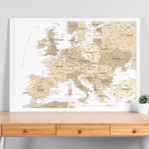 Peut inclure: Une carte encadrée de l'Europe, aux tons neutres beige et blanc. La carte affiche les pays avec leurs noms, entourée d'un cadre blanc. L'océan Atlantique est indiqué à gauche. Un petit pot de plante est visible.