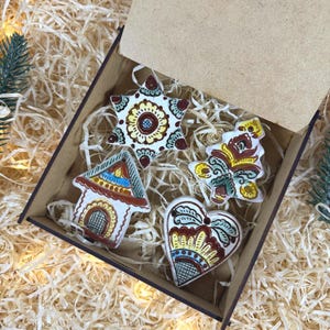May include: A set of four ceramic ornaments in a brown wooden box filled with wood shavings. The ornaments include a star, a house, a tree, and a heart, each hand-painted with intricate floral and geometric patterns in red, yellow, and blue.