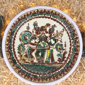 Puede incluir: Un plato de cerámica redondo con una ilustración pintada a mano de tres figuras con vestimenta tradicional tocando instrumentos musicales. El diseño utiliza una paleta de colores crema, marrón oscuro, verde y amarillo, con un borde decorativo.