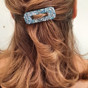 Peut inclure: Une femme avec de longs cheveux bruns portant un chemisier noir. Elle porte une pince à cheveux bleue à paillettes dans ses cheveux.