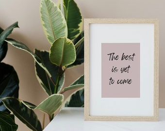 May include: A framed print with a pale pink background and the text "The best is yet to come" in black script. The frame is light wood and is sitting on a white surface next to a green plant.