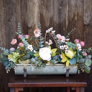 May include: A rectangular metal planter filled with artificial flowers and greenery. The arrangement features pink, white, and yellow blooms, with green foliage. The planter sits on a small wooden table against a rustic wooden backdrop.