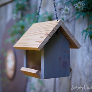 May include: A wooden bird feeder with a light brown roof and gray body hangs from a chain. The feeder has a tray for food and is set against a blurred background of greenery and a wooden fence. The words "Spring Creek Cottage" are at the bottom.