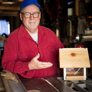 May include: A wooden birdhouse with a pitched roof and clear front panel. The birdhouse is light-colored and appears to be newly constructed. The birdhouse is on a table saw. The image is from Spring Creek Cottage.