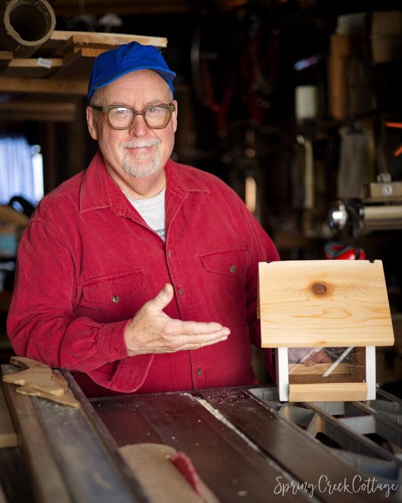 May include: A wooden birdhouse with a pitched roof and clear front panel. The birdhouse is light-colored and appears to be newly constructed. The birdhouse is on a table saw. The image is from Spring Creek Cottage.