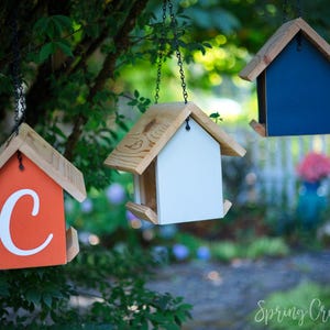 May include: Three hanging birdhouses with wooden roofs and various colored bodies: orange with a white "C", white, and navy blue. The birdhouses are suspended from chains and set against a blurred green background.