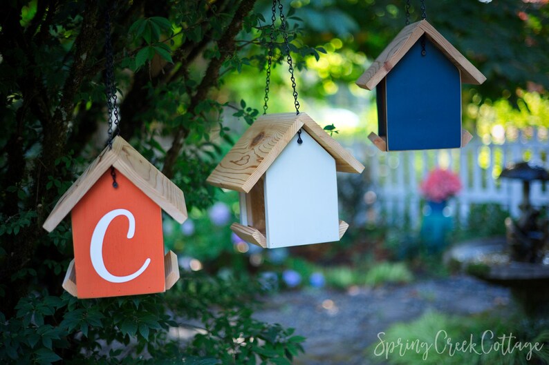 May include: Three hanging birdhouses with wooden roofs and various colored bodies: orange with a white "C", white, and navy blue. The birdhouses are suspended from chains and set against a blurred green background.