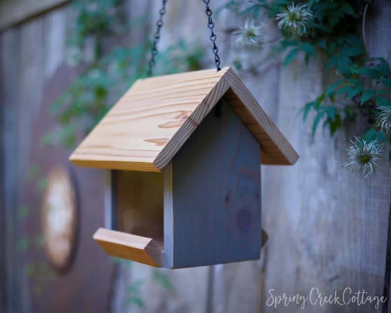 May include: A wooden bird feeder with a light brown roof and gray body hangs from a chain. The feeder has a tray for food and is set against a blurred background of greenery and a wooden fence. The words "Spring Creek Cottage" are at the bottom.