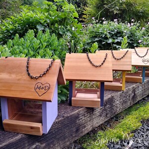 May include: A collection of wooden bird feeders with pitched roofs and chain hangers. Each feeder has a different colored base, including purple, blue, yellow, and red. Some feeders are engraved with a heart and initials. The feeders are displayed outdoors.