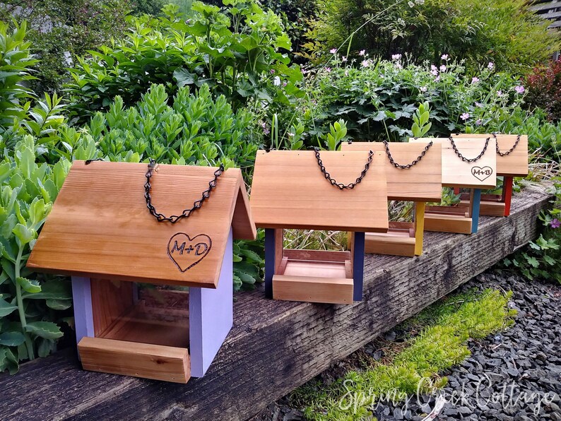May include: A collection of wooden bird feeders with pitched roofs and chain hangers. Each feeder has a different colored base, including purple, blue, yellow, and red. Some feeders are engraved with a heart and initials. The feeders are displayed outdoors.