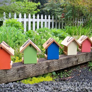 May include: A row of six colorful birdhouses, each with a different color: red, green, blue, cream, pink, and orange. The birdhouses are lined up on a wooden beam, with a white picket fence in the background.