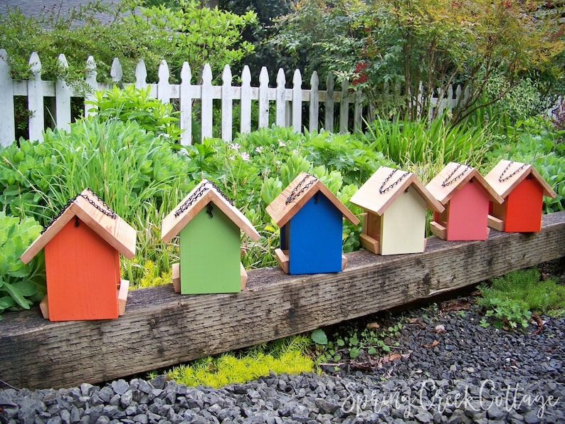 May include: A row of six colorful birdhouses, each with a different color: red, green, blue, cream, pink, and orange. The birdhouses are lined up on a wooden beam, with a white picket fence in the background.