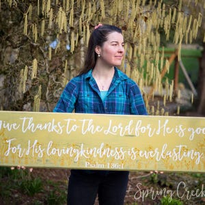 Może przedstawiać: Żółty drewniany znak z białym napisem: "Give thanks to the Lord for He is good, For His loving kindness is everlasting Psalm 136:1". Znak jest ozdobiony małymi żółtymi kwiatami.