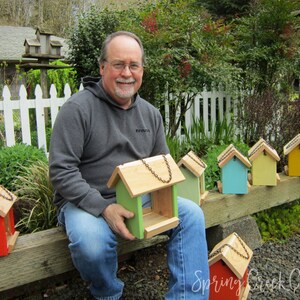 May include: A man holding a green and natural wood birdhouse with a chain for hanging. Several other colorful birdhouses in red, green, blue, and yellow are displayed on a wooden beam. The birdhouses have natural wood roofs.