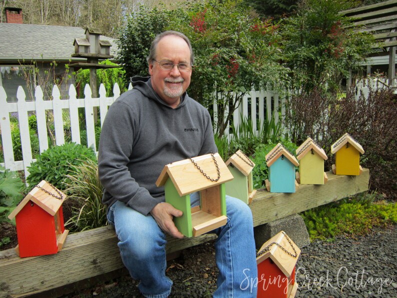May include: A man holding a green and natural wood birdhouse with a chain for hanging. Several other colorful birdhouses in red, green, blue, and yellow are displayed on a wooden beam. The birdhouses have natural wood roofs.