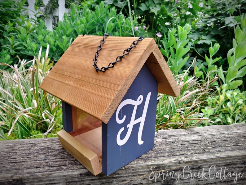 May include: Wooden bird feeder with a brown roof and navy blue body, featuring a white script letter "H". The feeder has a black chain for hanging and a wooden perch. The background shows green foliage and a white picket fence.
