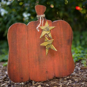 May include: A flat, orange-painted wooden pumpkin cutout with a distressed finish. The pumpkin features a stem wrapped in natural jute twine, from which two small, yellow-painted wooden stars are suspended by thin, dark metal wire.