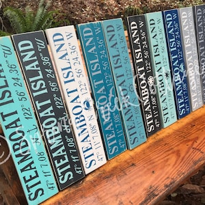 Puede incluir: Un conjunto de diez letreros de madera con las palabras "STEAMBOAT ISLAND" y coordenadas pintadas en ellos. Los letreros están pintados en varios tonos de azul, gris y blanco.