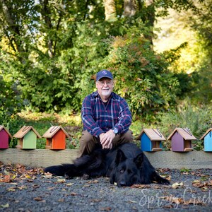 May include: A man wearing a plaid shirt and a baseball cap sits with a black dog in front of a row of colorful birdhouses. The birdhouses are painted in shades of blue, green, red, and purple.