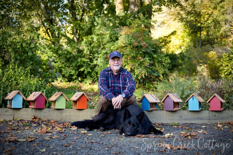 May include: A man wearing a plaid shirt and a navy blue cap sits on a black dog in front of a row of colorful birdhouses. The birdhouses are painted in various colors, including blue, red, green, and orange.