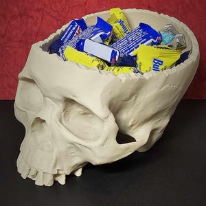 May include: A cream-colored skull-shaped container filled with assorted candy bars. The skull has detailed features and is set against a red and black background. The candy wrappers are blue, yellow, and white.