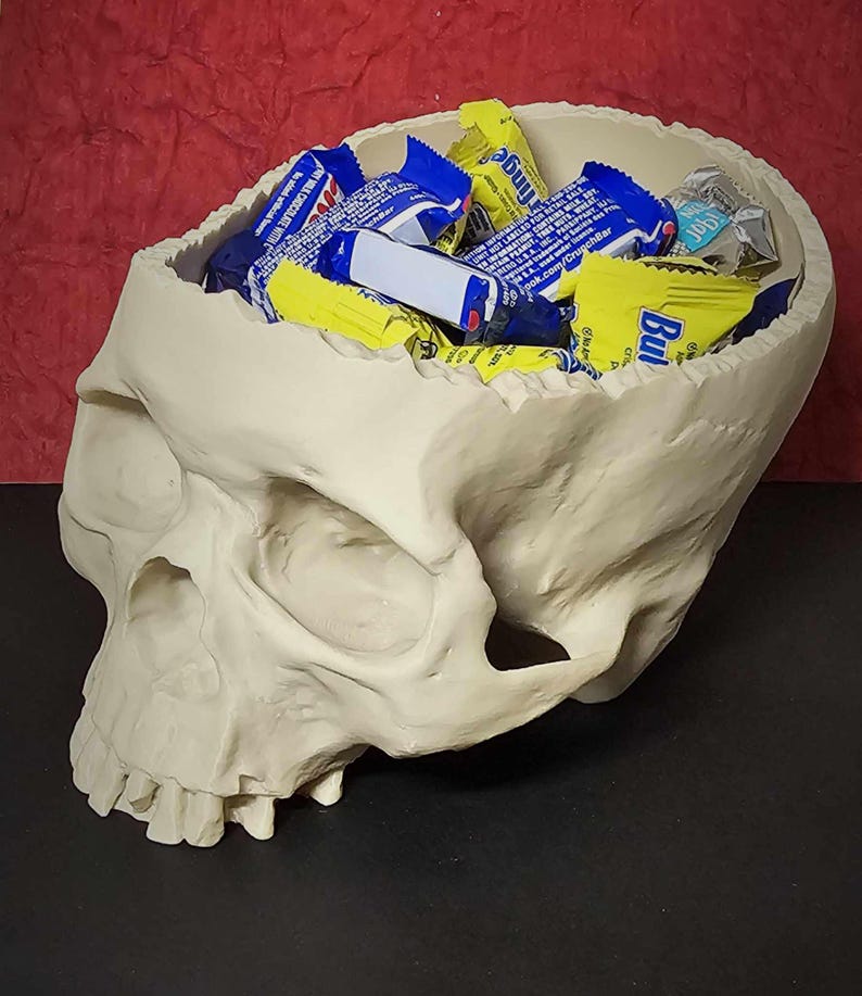 May include: A cream-colored skull-shaped container filled with assorted candy bars. The skull has detailed features and is set against a red and black background. The candy wrappers are blue, yellow, and white.