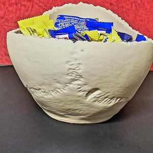 May include: A beige, skull-shaped bowl filled with yellow and blue candy wrappers. The bowl has a textured surface and is set against a red background. The bowl is a unique decorative piece.