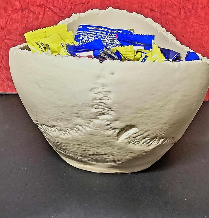 May include: A beige, skull-shaped bowl filled with yellow and blue candy wrappers. The bowl has a textured surface and is set against a red background. The bowl is a unique decorative piece.