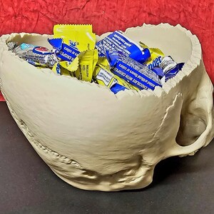 May include: A beige skull-shaped bowl filled with wrapped candy. The bowl has a rough texture and a handle on the side. The candy wrappers are yellow and blue.