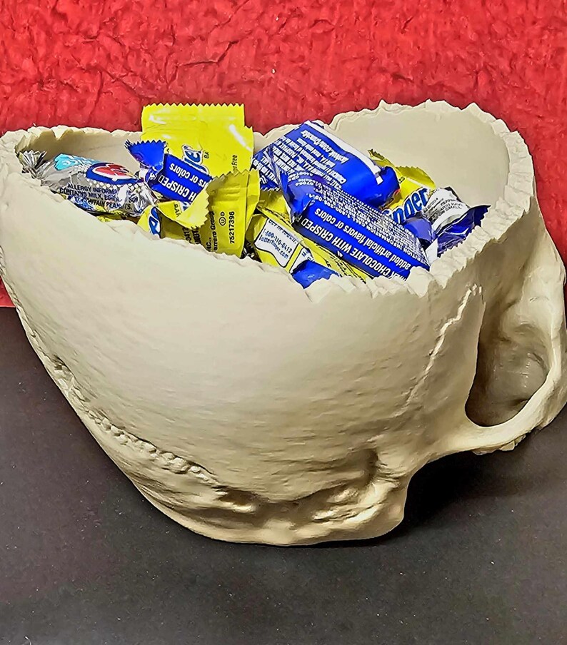 May include: A beige skull-shaped bowl filled with wrapped candy. The bowl has a rough texture and a handle on the side. The candy wrappers are yellow and blue.