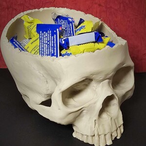 May include: A cream-colored skull-shaped container filled with individually wrapped candies. The candies are in blue and yellow wrappers. The skull has detailed features and is resting on a dark surface.