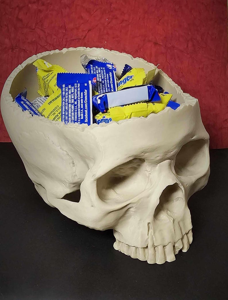 May include: A cream-colored skull-shaped container filled with individually wrapped candies. The candies are in blue and yellow wrappers. The skull has detailed features and is resting on a dark surface.