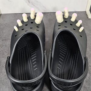 May include: A pair of black Crocs with attached toe-shaped accessories. The toes are beige with pink nail polish. The shoes are on a gray surface.