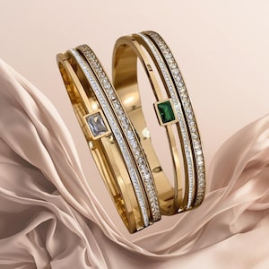 May include: Two gold-coloured bangles with sparkling clear and white gemstones. One bangle features a square, light blue gemstone, while the other has a square, green gemstone. The bangles are displayed on a soft, peach-coloured fabric.