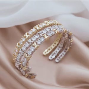 May include: Three open cuff bracelets. One is gold-toned, one is silver-toned, and the third is a mix of gold and silver. Each bracelet features a row of clear, round gemstones and an X-shaped design. The bracelets are displayed on a satin surface.