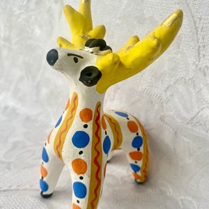 May include: A white ceramic deer figurine with yellow antlers, orange and blue polka dots, and wavy gold lines. The deer has a black face with a small black nose and black eyes.